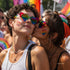 Pride You! | Occhiali da Sole a Cuore Arcobaleno (Love Celebration Special Pick) WYNIE indossati da una coppia di donne che si baciano sulla guancia durante una manifestazione del PRIDE, celebrando l'amore e la libertà di esperimersi