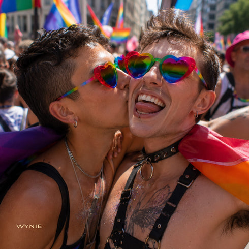 Pride You! | Occhiali da Sole a Cuore Arcobaleno (Love Celebration Special Pick) WYNIE indossati da una coppia di uomini che si baciano sulla guancia durante una manifestazione del PRIDE, celebrando l'amore e la libertà di esperimersi