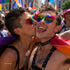 Pride You! | Occhiali da Sole a Cuore Arcobaleno (Love Celebration Special Pick) WYNIE indossati da una coppia di uomini che si baciano sulla guancia durante una manifestazione del PRIDE, celebrando l'amore e la libertà di esperimersi