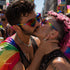 Pride You! | Occhiali da Sole a Cuore Arcobaleno (Love Celebration Special Pick) WYNIE indossati da una coppia queer che si bacia sulla guancia durante una manifestazione del PRIDE, celebrando l'amore e la libertà di esperimersi