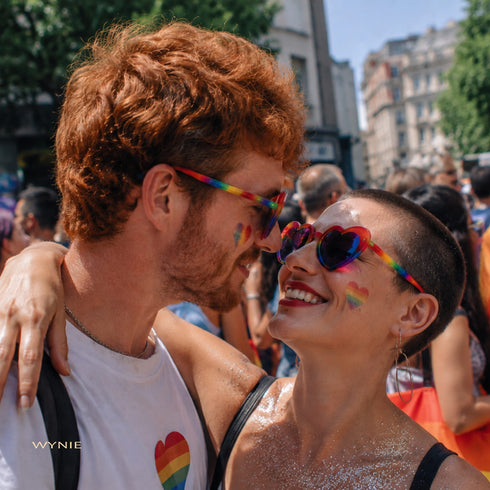 Pride You! | Occhiali da Sole a Cuore Arcobaleno (Love Celebration Special Pick) WYNIE indossati da una coppia uomo e donna che si guardano negli occhi durante una manifestazione del PRIDE, celebrando l'amore e la libertà di esperimersi